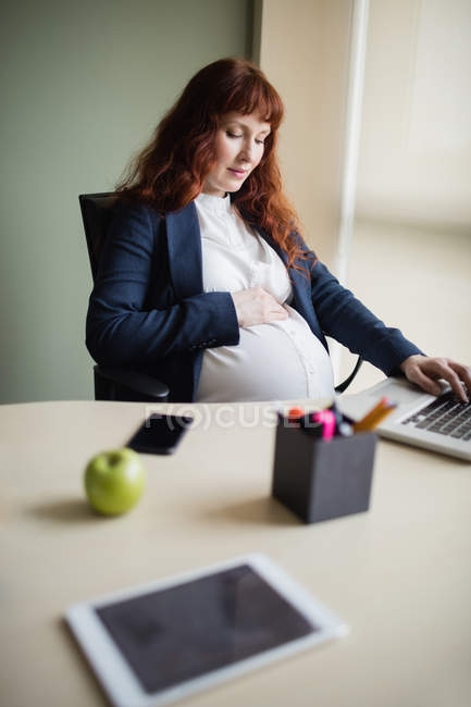 Вагітна бізнес-леді торкається живота, використовуючи ноутбук в офісі — стокове фото