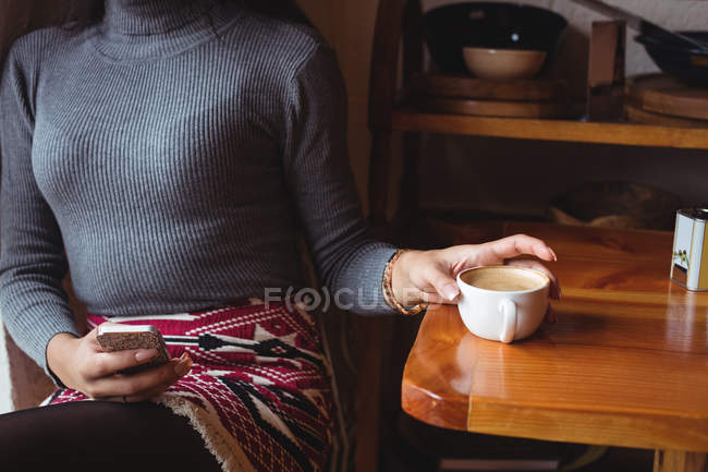 Середина жінки, використовуючи мобільний телефон, маючи чашку кави в кафе — стокове фото