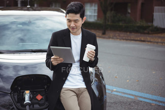 Man using digital tablet while charging car at electric vehicle charging station — Stock Photo