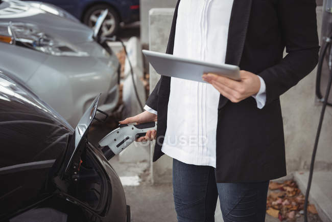 Seção média da mulher usando o tablet digital ao carregar o carro elétrico na estação de carregamento do veículo — Fotografia de Stock