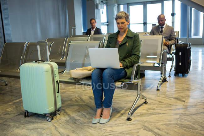Mujer con equipaje usando portátil en la sala de espera en la terminal del aeropuerto - foto de stock