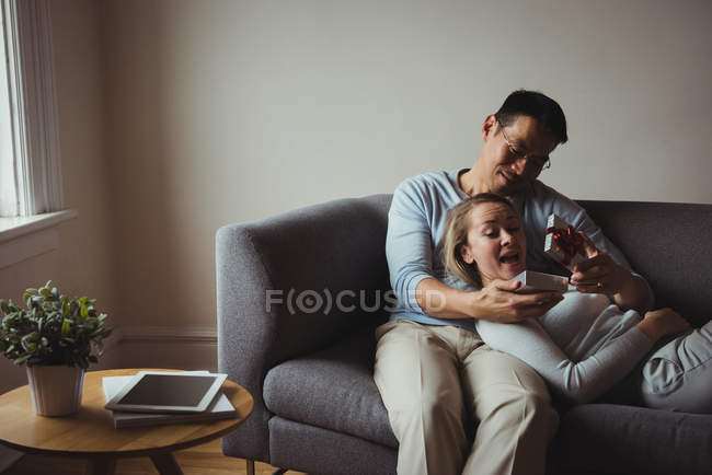 Homem dando presente surpresa para sua mulher no sofá em casa — Fotografia de Stock