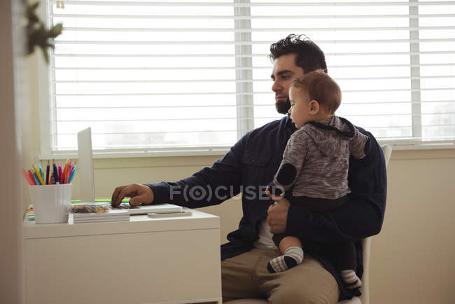 Pai segurando seu bebê enquanto usa laptop na mesa em casa — Fotografia de Stock