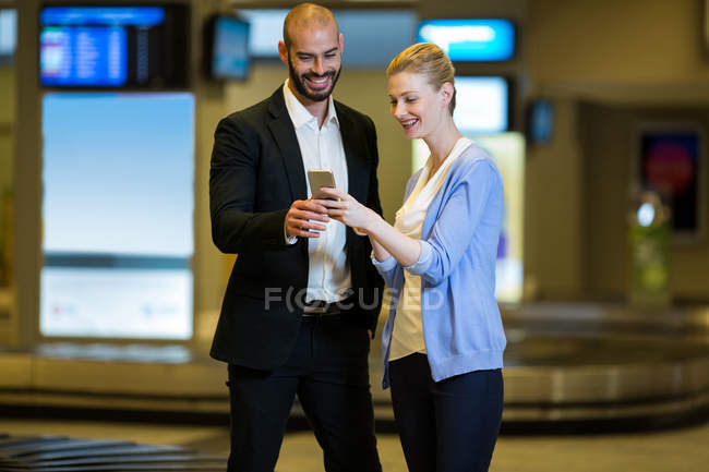 Улыбающаяся пара смотрит на мобильный телефон в зоне ожидания в терминале аэропорта — стоковое фото