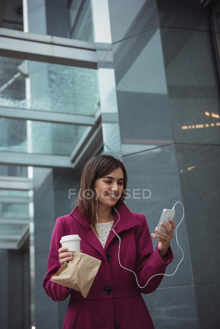 Donna d'affari in possesso di tazza di caffè usa e getta e pacchetto durante l'ascolto di musica vicino edificio per uffici — Foto stock