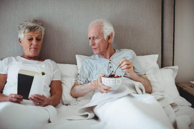 Älterer Mann frühstückt, während Frau ein Buch auf Bett im Schlafzimmer liest — Stockfoto