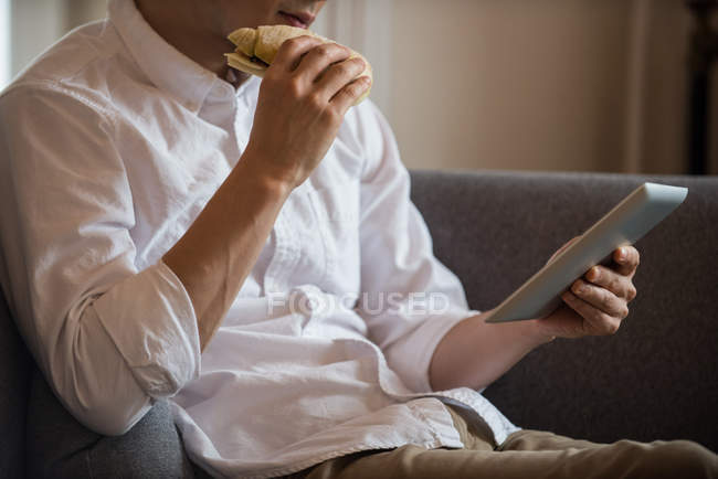 Man using digital tablet while having sandwich at home — Stock Photo