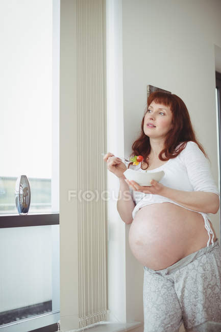 Donna incinta premurosa che ha insalata vicino alla finestra a casa — Foto stock