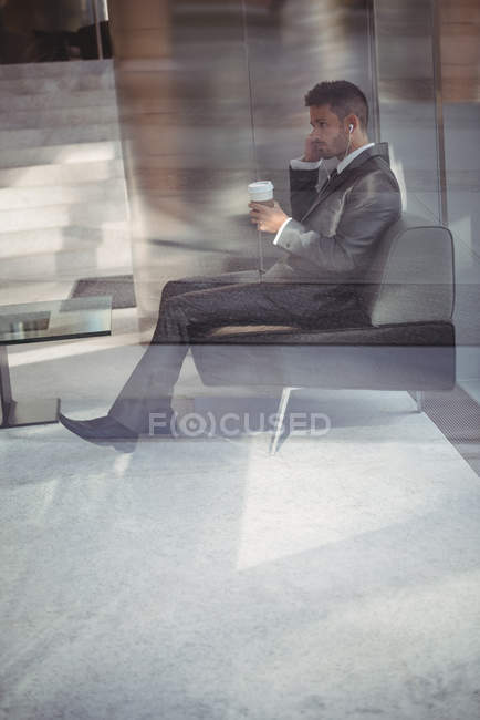 Бизнесмен держит одноразовый кофейный стаканчик и слушает музыку, сидя на диване в офисных помещениях — стоковое фото