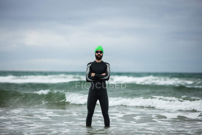 Athlete in wet suit standing with arms crossed in sea on beach — Stock Photo