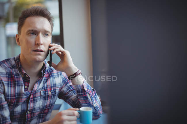 Jovem falando no celular enquanto toma café no refeitório — Fotografia de Stock