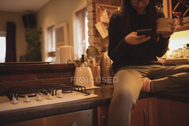 Woman using mobile phone while having coffee in kitchen — Stock Photo