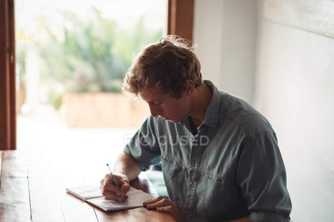 Чоловік сидить за столом, пишучи на блокноті вдома — стокове фото