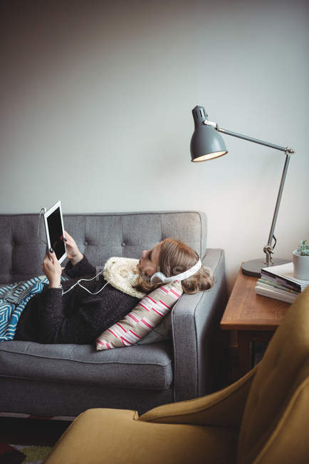 Woman lying on sofa listening to music on digital tablet at home — Stock Photo