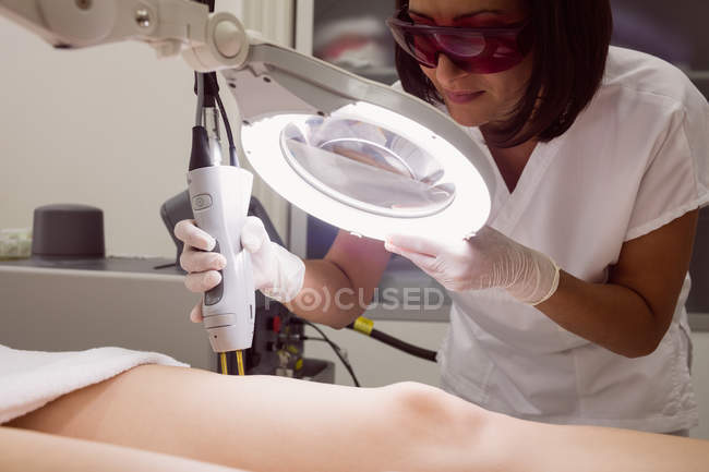 Крупним планом лікар, що виконує лазерну епіляцію на шкірі стегна пацієнта в клініці — стокове фото