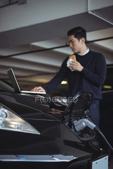 Hombre usando el ordenador portátil mientras carga coche eléctrico en el garaje - foto de stock