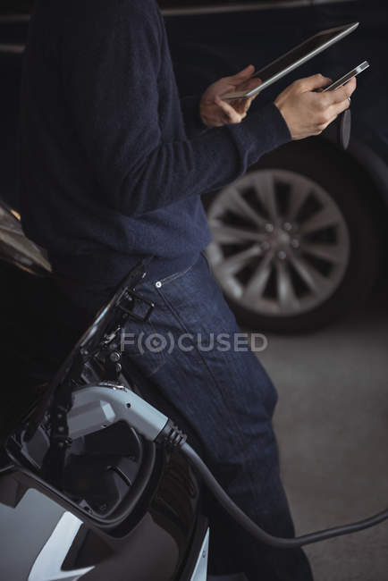 Mid section of man using digital tablet and mobile phone while charging electric car in garage — Stock Photo