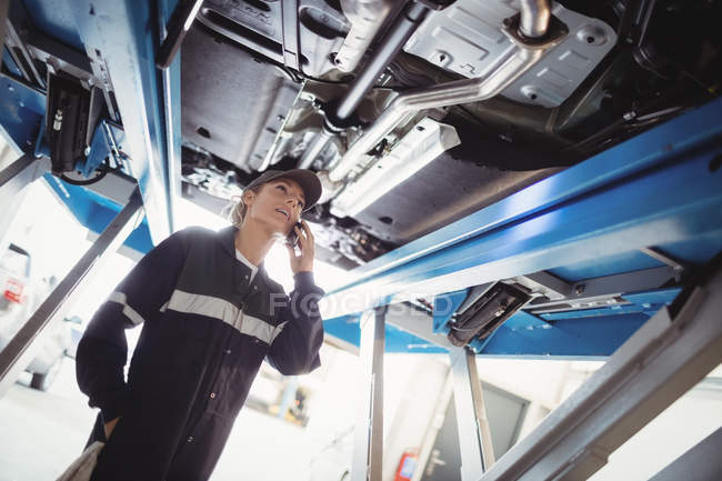 Женщина-механик разговаривает по мобильному телефону под машиной в ремонтном гараже — стоковое фото