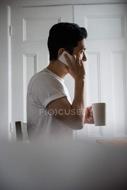 Homem falando no celular enquanto toma uma xícara de café em casa — Fotografia de Stock