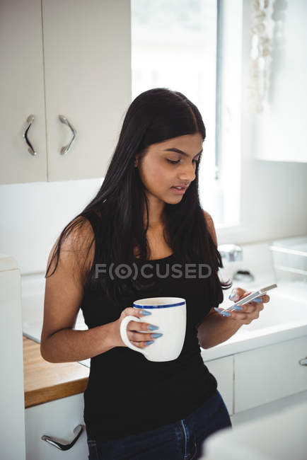 Woman using mobile phone while holding cup of coffee in kitchen at home — Stock Photo
