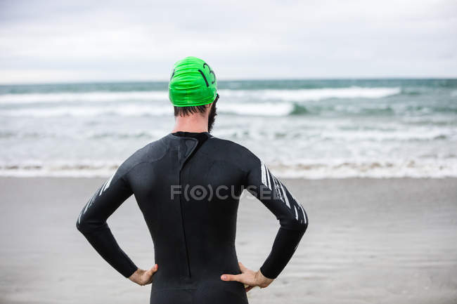Visão traseira do atleta de terno molhado de pé com as mãos na cintura na praia — Fotografia de Stock