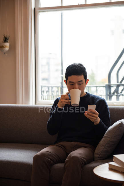 Uomo che utilizza il telefono cellulare mentre prende una tazza di caffè a casa — Foto stock