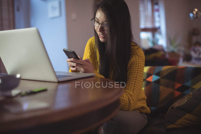 Mujer usando teléfono móvil con portátil en la mesa en casa - foto de stock