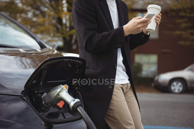 Средняя часть человека с помощью мобильного телефона во время зарядки автомобиля на электростанции зарядки автомобиля — стоковое фото