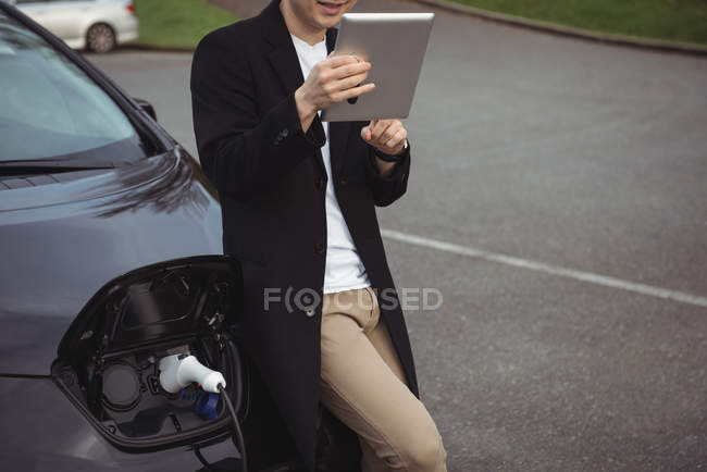 Человек с цифровым планшетом во время зарядки электромобиля на электростанции зарядки автомобиля — стоковое фото