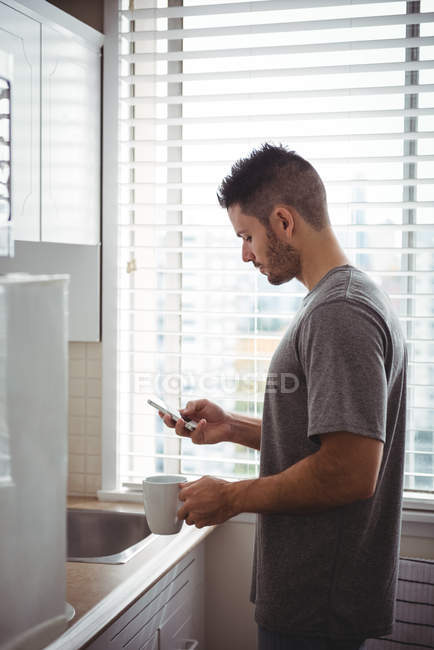 Homme utilisant son téléphone portable tout en tenant une tasse de café dans la cuisine à la maison — Photo de stock