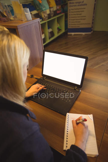 Frau benutzt Laptop und schreibt im Büro auf Notizblock — Stockfoto