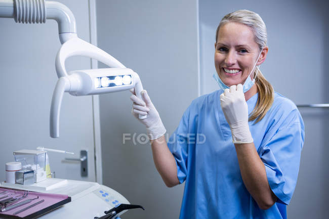 Assistant dentaire souriant à la caméra à côté de la lumière à la clinique dentaire — Photo de stock