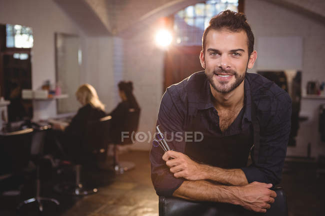 Retrato de sorrindo cabeleireiro masculino inclinado na cadeira no salão — Fotografia de Stock