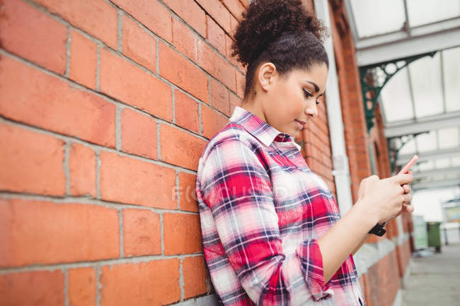 Vista lateral da mulher usando telefone enquanto se inclina na parede de tijolo — Fotografia de Stock