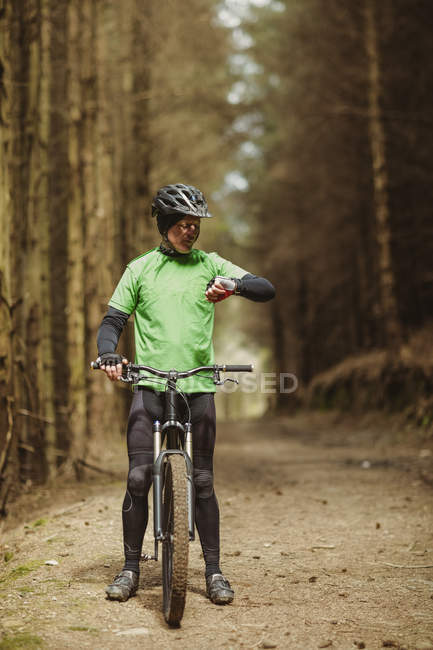 Vista frontal do ciclista de montanha verificando o tempo enquanto andava na floresta — Fotografia de Stock