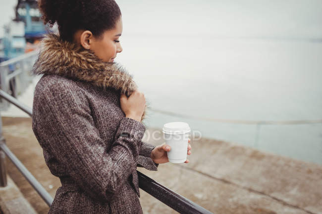 Side view of woman holding disposable coffee cup while standing by railing — Stock Photo