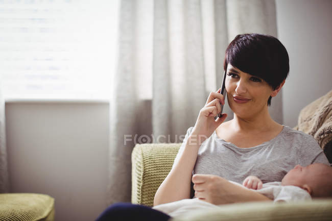 Mother talking by smartphone while baby sleeping in her arm at living room — Stock Photo