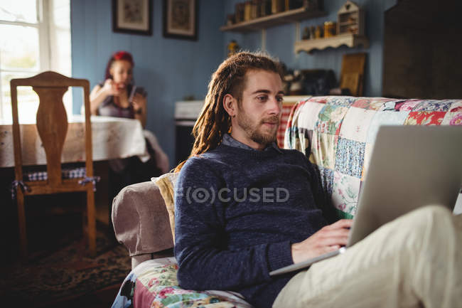 Hipster homem usando laptop enquanto mulher sentada no fundo em casa — Fotografia de Stock