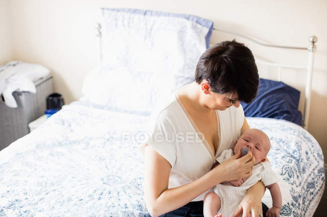 Mãe colocando manequim em sua boca bebê na cama no quarto em casa — Fotografia de Stock