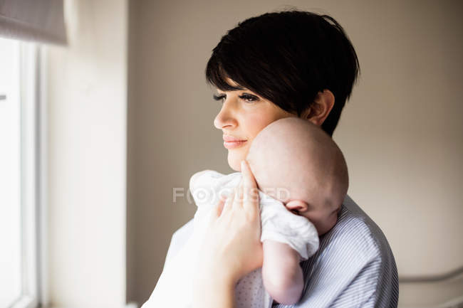 Mãe de pé perto da janela e segurando seu bebê em casa — Fotografia de Stock