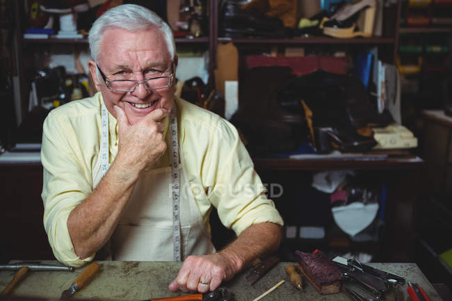 Retrato del zapatero sentado en el taller con la mano en la barbilla - foto de stock
