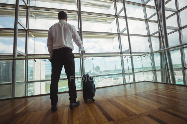 Vue arrière d'un homme d'affaires avec des bagages regardant par une fenêtre vitrée à l'aéroport — Photo de stock