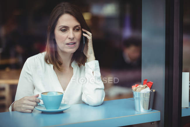 Woman holding coffee cup and talking on mobile phone in cafeteria — Stock Photo