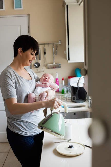Мать держит своего малыша, наливая кофе из электрического чайника на кухне — стоковое фото