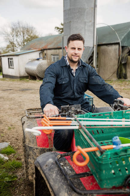 Portrait of smiling young farmer riding quadbike on field against barn — Stock Photo