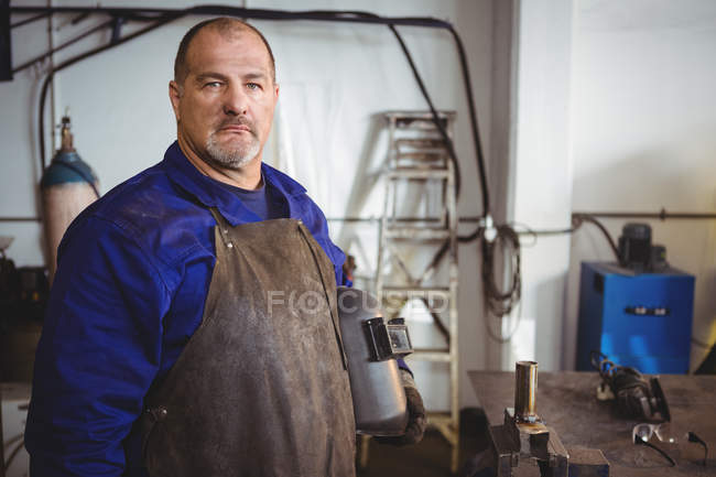 Retrato del soldador que sostiene el casco de soldadura en el taller - foto de stock