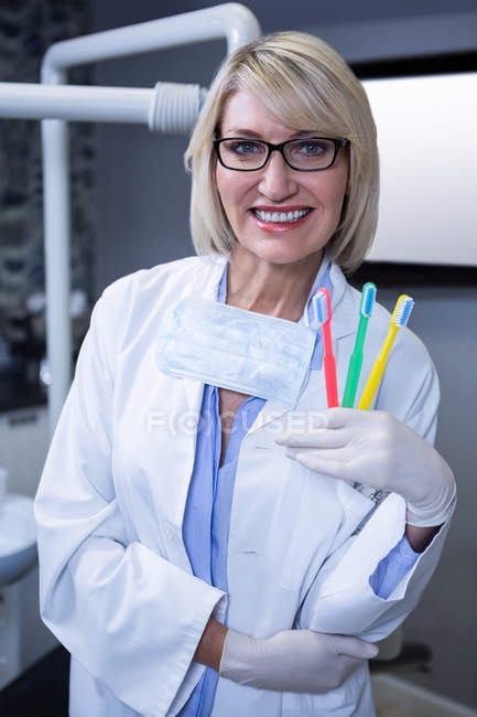 Retrato del dentista sonriente sosteniendo tres cepillos de dientes en la clínica dental - foto de stock
