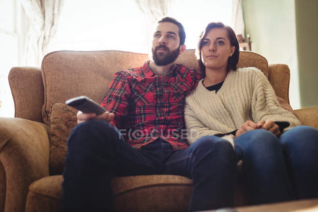Couple sitting on sofa with remote control while watching television in living room at home — Stock Photo
