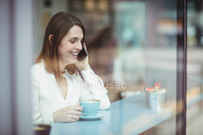 Mulher segurando xícara de café e falando no celular na cafetaria — Fotografia de Stock