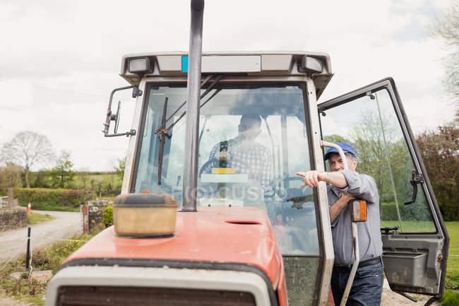 Работник фермы указывает на поле, стоя на тракторе — стоковое фото